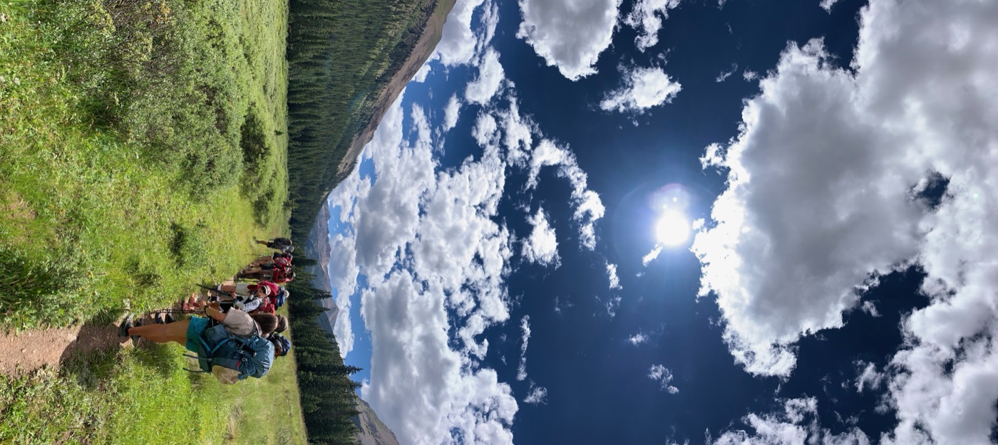 A line of backpackers hiking single-file up a green mountain valley trail beneath a vast, cloud-filled sky