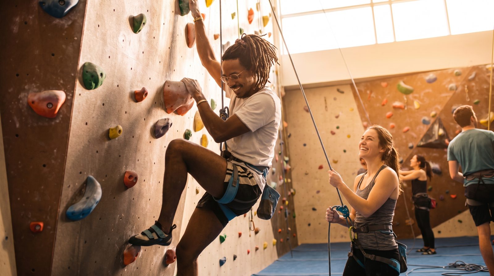 A climber laughs mid-route while a friend belays him from below in a warm, sunlit climbing gym